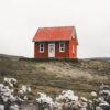 A small red house with a pitched roof stands alone on a grassy landscape, with white wildflowers in the foreground under an overcast sky. Nature and landscape poster print available in A1 to A4 sizes.