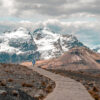 A lone person walks along a mountain path, paved and winding toward snow-capped mountains under a cloudy sky. Nature and landscape poster print available in A1 to A4 sizes.