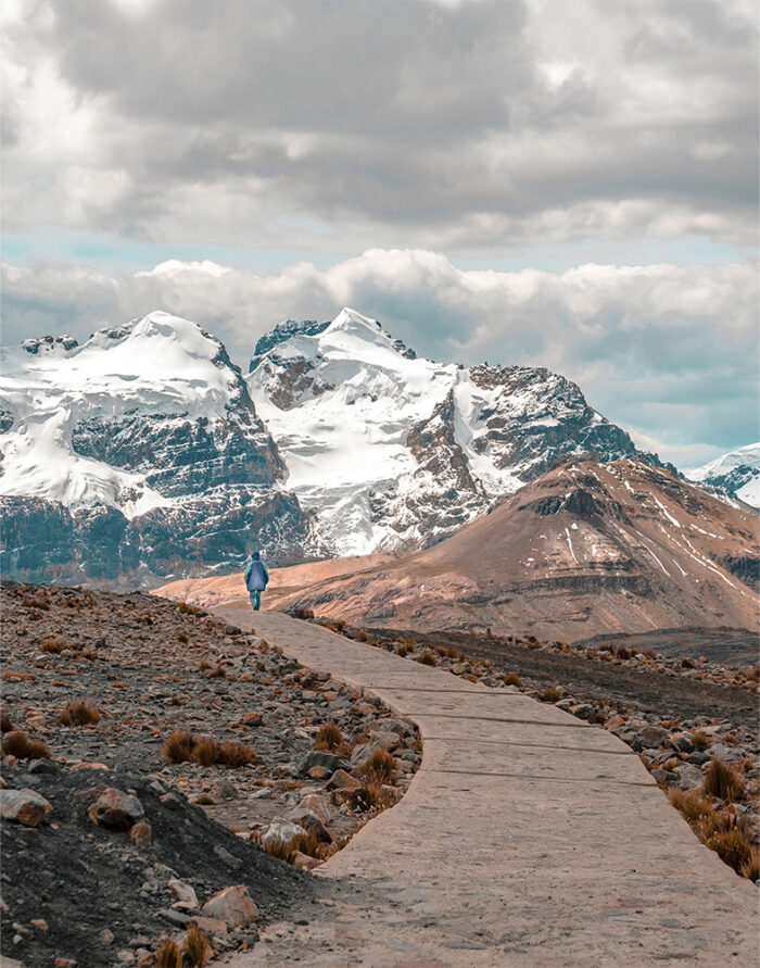 product-image-11-14 A lone person walks along a mountain path, paved and winding toward snow-capped mountains under a cloudy sky. Nature and landscape poster print available in A1 to A4 sizes.