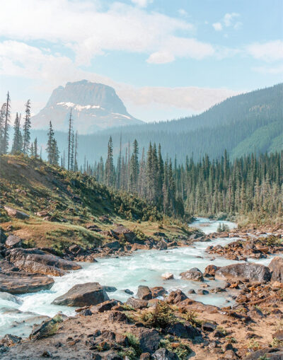 A mountain river winds through a forested valley with pine trees and rugged rocks, set against mountains in the background beneath a partly cloudy sky. Nature and landscape poster print available in A1 to A4 sizes.