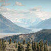 Snow-capped mountains rise in the background, creating a stunning mountain landscape with dense evergreen forests and a river valley in the foreground under a partly cloudy sky. Nature and landscape poster print available in A1 to A4 sizes.