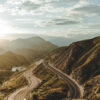 A winding mountain road curves through green hills with a few cars driving, mountains and a cloudy sky in the background. Nature and landscape poster print available in A1 to A4 sizes.