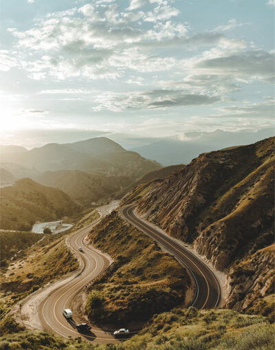 A winding mountain road curves through green hills with a few cars driving, mountains and a cloudy sky in the background. Nature and landscape poster print available in A1 to A4 sizes.