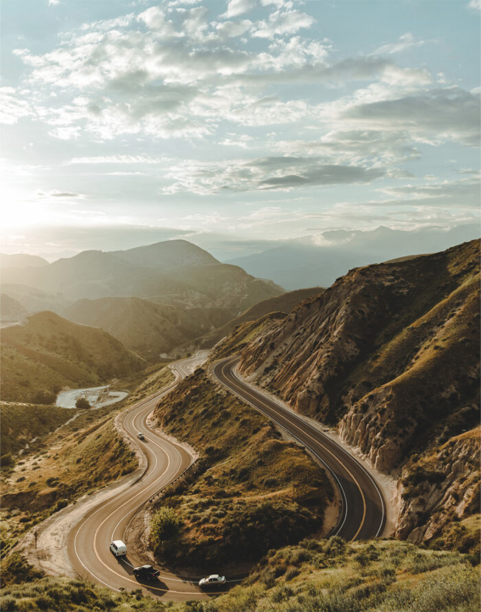 product-image-11-14 A winding mountain road curves through green hills with a few cars driving, mountains and a cloudy sky in the background. Nature and landscape poster print available in A1 to A4 sizes.