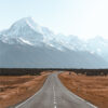 A straight mountain road leads toward snow-capped mountains under a clear sky, surrounded by dry grassland and distant trees. Nature and landscape poster print available in A1 to A4 sizes.