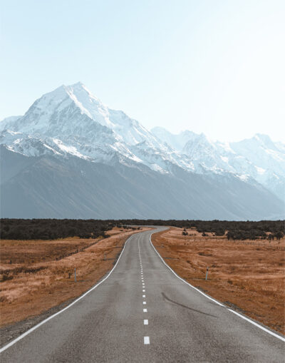 A straight mountain road leads toward snow-capped mountains under a clear sky, surrounded by dry grassland and distant trees. Nature and landscape poster print available in A1 to A4 sizes.