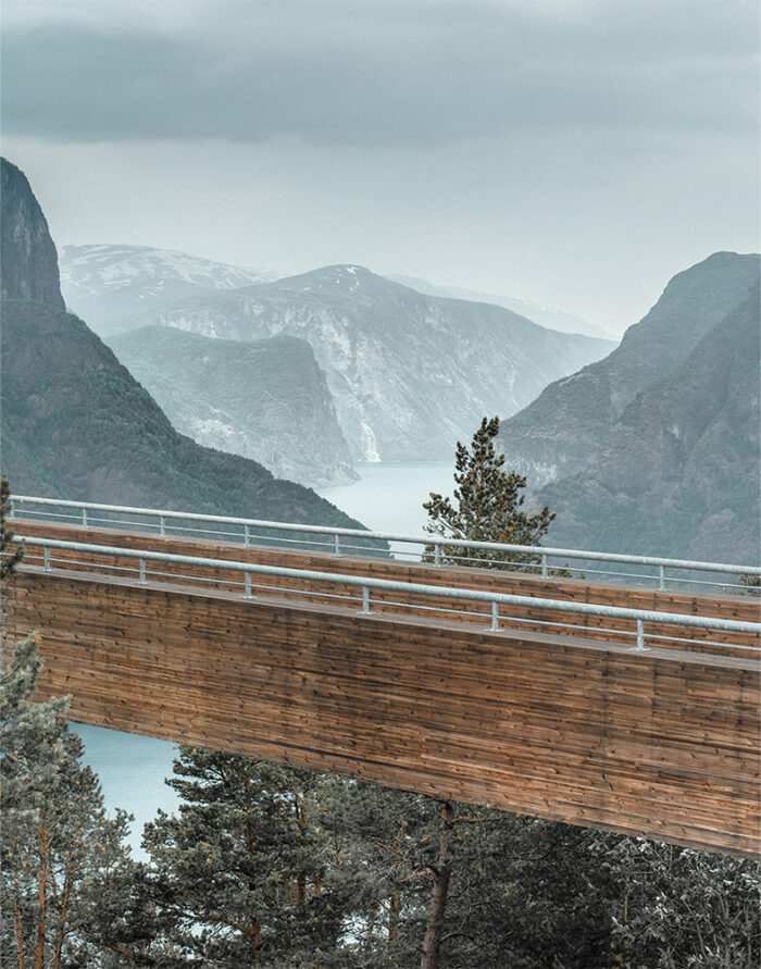 product-image-11-14 A wooden Mountain Walkway viewing platform extends over trees with a scenic backdrop of fjords and mountains under a cloudy sky. Nature and landscape poster print available in A1 to A4 sizes.