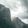 Tall, rugged mountain cliffs rise beneath a cloudy sky along a dramatic mountain ridge, with patches of snow and a small waterfall cascading down the rocky surface. Nature and landscape poster print available in A1 to A4 sizes.