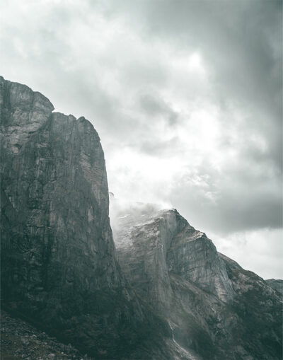 Tall, rugged mountain cliffs rise beneath a cloudy sky along a dramatic mountain ridge, with patches of snow and a small waterfall cascading down the rocky surface. Nature and landscape poster print available in A1 to A4 sizes.