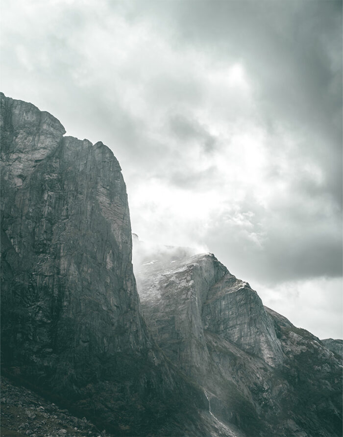product-image-11-14 Tall, rugged mountain cliffs rise beneath a cloudy sky along a dramatic mountain ridge, with patches of snow and a small waterfall cascading down the rocky surface. Nature and landscape poster print available in A1 to A4 sizes.