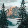 A tall, rocky mountain peak is illuminated by sunlight, framed by pine trees in the foreground, with clouds or mist covering the lower landscape. Nature and landscape poster print available in A1 to A4 sizes.