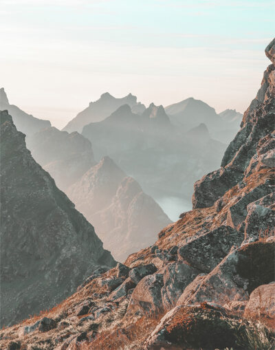 Rocky mountain peaks layered in mist, with rugged cliffs in the foreground and a pale sky above, create a breathtaking Mountain View. Nature and landscape poster print available in A1 to A4 sizes.