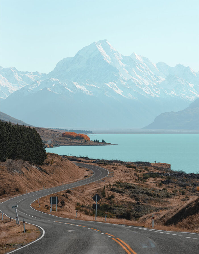 product-image-11-14 A winding Mountain Road leads toward a large, snow-covered mountain beside a serene lake, with sparse trees and dry grass in the foreground. Nature and landscape poster print available in A1 to A4 sizes.