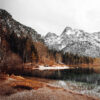 A calm mountain lake with a rocky shore, bordered by trees and overlooked by snow-capped mountains under a cloudy sky. Nature and landscape poster print available in A1 to A4 sizes.