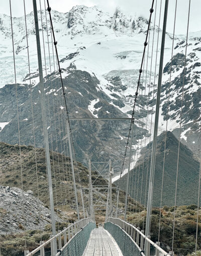 A mountain bridge stretches across rugged terrain, leading towards snow-covered peaks beneath a cloudy sky. Nature and landscape poster print available in A1 to A4 sizes.