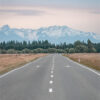A straight empty Mountain Road leads toward distant snow-capped mountains, bordered by fields and trees under a cloudy sky. Nature and landscape poster print available in A1 to A4 sizes.