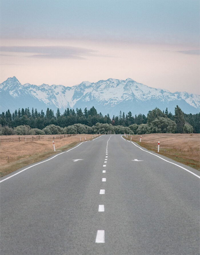 product-image-11-14 A straight empty Mountain Road leads toward distant snow-capped mountains, bordered by fields and trees under a cloudy sky. Nature and landscape poster print available in A1 to A4 sizes.