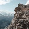 A person stands on a rocky mountain peak overlooking a breathtaking Mountain View, with snow-capped mountains and clouds stretching across the distant valley. Nature and landscape poster print available in A1 to A4 sizes.