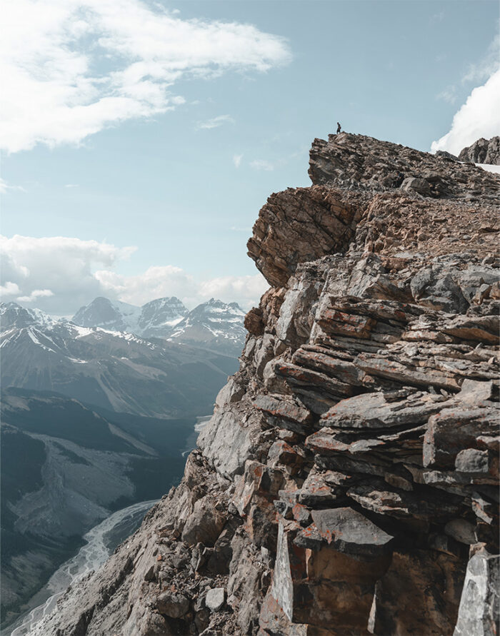 product-image-11-14 A person stands on a rocky mountain peak overlooking a breathtaking Mountain View, with snow-capped mountains and clouds stretching across the distant valley. Nature and landscape poster print available in A1 to A4 sizes.