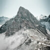 Rocky mountain peak rises above clouds with patches of snow on its slopes; part of a majestic mountain range, with other peaks visible in the background under a pale sky. Nature and landscape poster print available in A1 to A4 sizes.