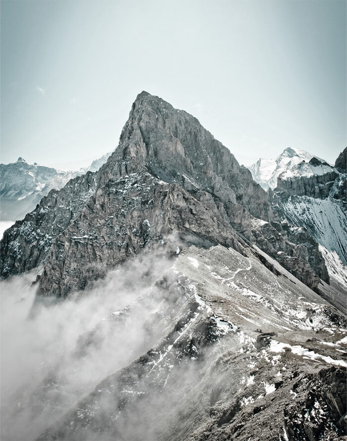 product-image-11-14 Rocky mountain peak rises above clouds with patches of snow on its slopes; part of a majestic mountain range, with other peaks visible in the background under a pale sky. Nature and landscape poster print available in A1 to A4 sizes.