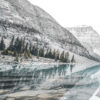 A calm mountain lake reflects pine trees and rocky mountains on a clear day, with an overcast sky and muted colours. Nature and landscape poster print available in A1 to A4 sizes.