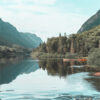A calm mountain lake surrounded by forested mountains, with two orange canoes docked along the shore under a partly cloudy sky. Nature and landscape poster print available in A1 to A4 sizes.