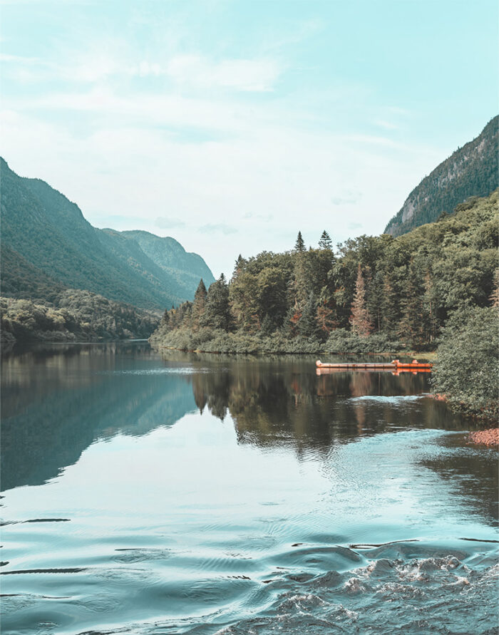 product-image-11-14 A calm mountain lake surrounded by forested mountains, with two orange canoes docked along the shore under a partly cloudy sky. Nature and landscape poster print available in A1 to A4 sizes.