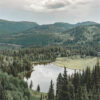 A small lake is surrounded by dense evergreen trees and rolling green hills under a cloudy sky. Nature and landscape poster print available in A1 to A4 sizes.