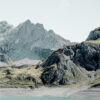 A turquoise lake stretches along a rugged shoreline, bordered by rocky hills and steep mountain slopes under a clear sky. Nature and landscape poster print available in A1 to A4 sizes.