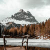 A rustic wooden dock overlooks a partially frozen Winter Mountain Lake, with snow-capped peaks and pine trees in the background under a cloudy sky. Nature and landscape poster print available in A1 to A4 sizes.