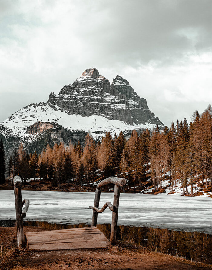 product-image-11-14 A rustic wooden dock overlooks a partially frozen Winter Mountain Lake, with snow-capped peaks and pine trees in the background under a cloudy sky. Nature and landscape poster print available in A1 to A4 sizes.