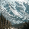 A paved road winds through a forest toward tall, snow-covered mountains under a cloudy sky. Nature and landscape poster print available in A1 to A4 sizes.