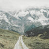 A gravel mountain trail winds through green hills toward tall, snow-capped mountains partially covered by clouds under an overcast sky. Nature and landscape poster print available in A1 to A4 sizes.