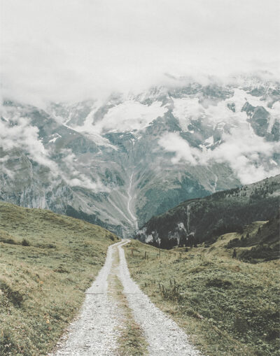 A gravel mountain trail winds through green hills toward tall, snow-capped mountains partially covered by clouds under an overcast sky. Nature and landscape poster print available in A1 to A4 sizes.