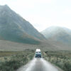 A white van drives along a Mountain Road through a green valley, with large mountains towering in the background under a cloudy sky. Nature and landscape poster print available in A1 to A4 sizes.