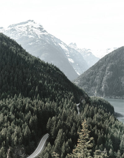 A winding stretch of Road runs through dense pine forests, with snow-capped mountains and a tranquil lake in the background. Nature and landscape poster print available in A1 to A4 sizes.