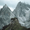 A person wearing a yellow jacket stands on a grassy mountain ridge, with steep, rocky mountains rising in the background under a cloudy sky. Nature and landscape poster print available in A1 to A4 sizes.