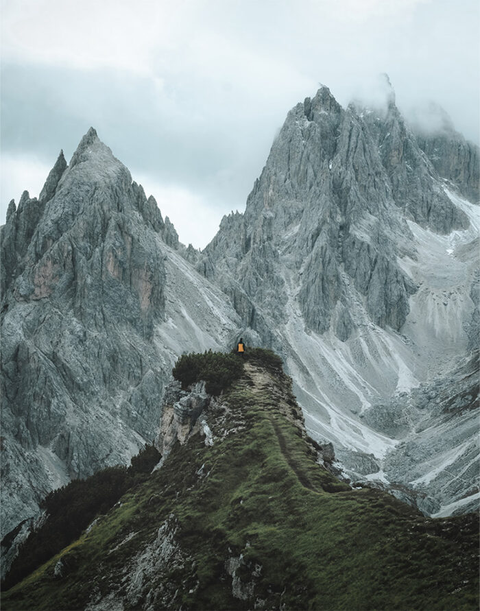 product-image-11-14 A person wearing a yellow jacket stands on a grassy mountain ridge, with steep, rocky mountains rising in the background under a cloudy sky. Nature and landscape poster print available in A1 to A4 sizes.