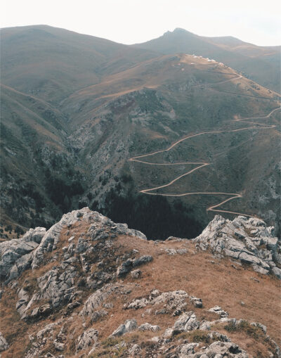 Rocky terrain in the foreground with a winding hiking trail leading through hilly, grassy mountains under a cloudy sky. Nature and landscape poster print available in A1 to A4 sizes.
