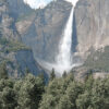A tall mountain waterfall cascades down a rocky cliff surrounded by trees, with a clear blue sky above. Nature and landscape poster print available in A1 to A4 sizes.