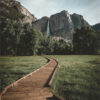 A wooden boardwalk serves as a scenic hiking trail through a grassy meadow toward a waterfall cascading down a rocky mountain in the distance, surrounded by trees. Nature and landscape poster print available in A1 to A4 sizes.