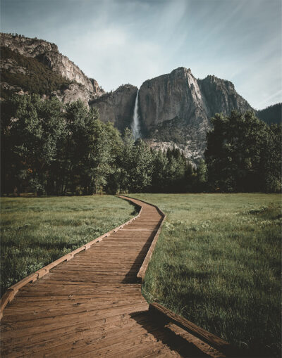 A wooden boardwalk serves as a scenic hiking trail through a grassy meadow toward a waterfall cascading down a rocky mountain in the distance, surrounded by trees. Nature and landscape poster print available in A1 to A4 sizes.