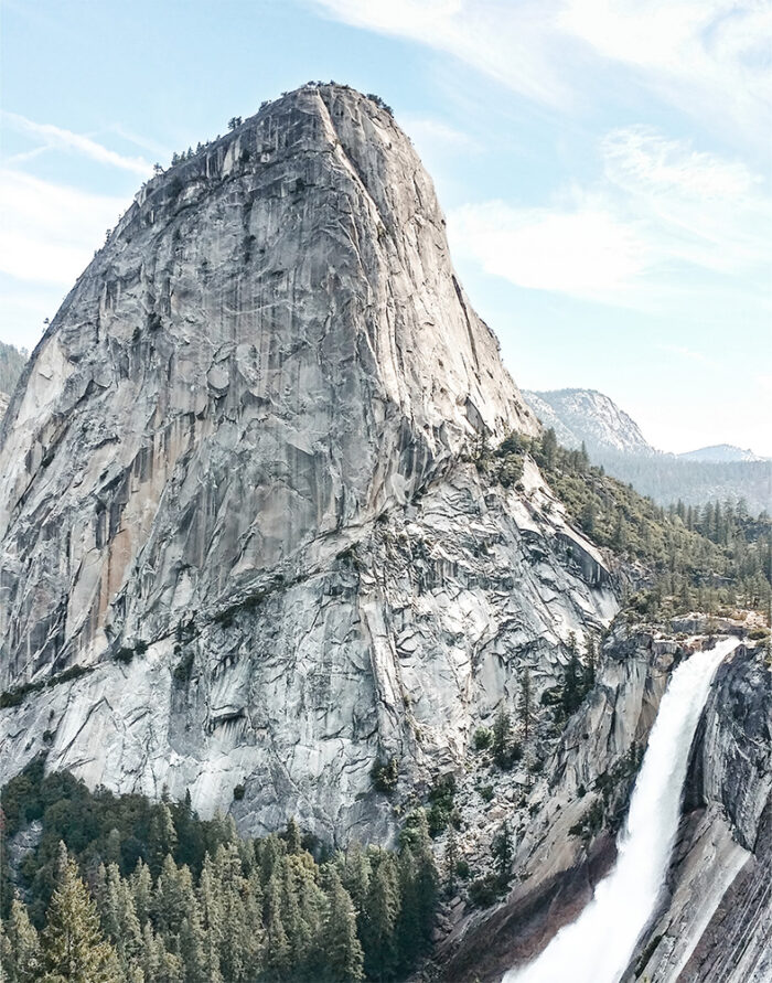 A steep granite mountain rises beside a cascading waterfall, surrounded by pine trees under a partly cloudy sky. Nature and landscape poster print available in A1 to A4 sizes.