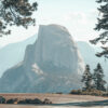 A large granite dome, known as Half Dome, rises behind pine trees and a Mountain Road, winding through Yosemite National Park on a clear day. Nature and landscape poster print available in A1 to A4 sizes.