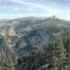 Mountain View with dense pine forests, rocky cliffs, and a distant waterfall under a partly cloudy sky. Nature and landscape poster print available in A1 to A4 sizes.