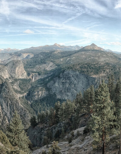 Mountain View with dense pine forests, rocky cliffs, and a distant waterfall under a partly cloudy sky. Nature and landscape poster print available in A1 to A4 sizes.