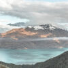 Clouds drift over a snow-capped peak above forested hills and turquoise waters of a Mountain Lake, where a small boat glides across, leaving a gentle wake behind. Nature and landscape poster print available in A1 to A4 sizes.