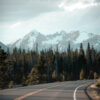 A paved mountain road curves through a forest with tall pine trees, leading toward distant snow-capped mountains under a cloudy sky. Nature and landscape poster print available in A1 to A4 sizes.