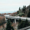 A curved Mountain Road winds through a forested, hilly landscape with autumn foliage and evergreen trees beneath an overcast sky. Nature and landscape poster print available in A1 to A4 sizes.